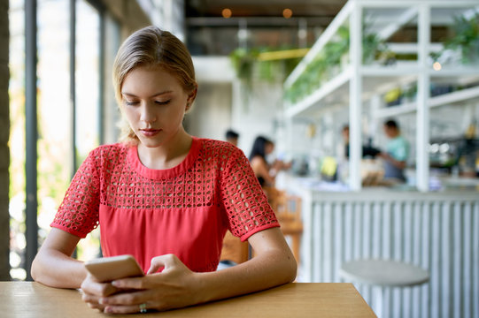 Lifestyle shot of young blonde caucasian natural millennial woman typing on cellphone at a modern tropical and bright restaurant with plants