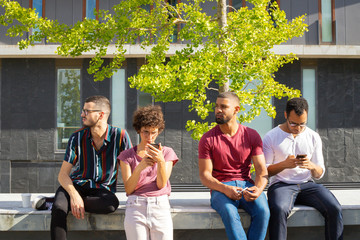 Group of friends consulting mobile app outdoors. Men and woman standing outside, sitting on parapet and using smartphones. Networking concept