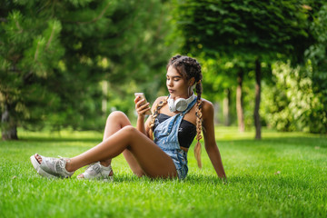 Image of young african girl on grass outdoors in park. She looks social networks at her phone.