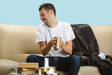 Young man suffering from hausehold dust or seasonal allergy. Sneezing in the napkin and sitting...