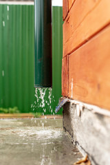 Rain water flows from a drainpipe to a concrete foundation, a wooden house, against a green fence.