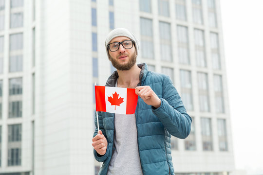 Portrait of a young man with a beard in a jacket and hat with the flag of Canada against the backdrop of the city. Study abroad and travel. Learn English language.