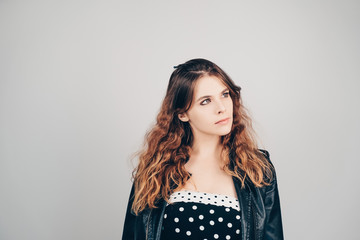 Pretty young woman wearing black leather jacket and retro dress looking aside, grey background. Portrait of beautiful european girl  with curly wavy ombre hair. 