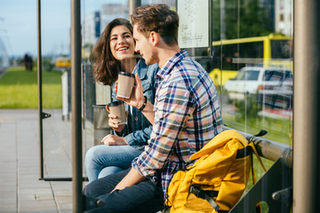 Positive relax couple traveler drinking coffee outdoor at stop while waiting tram, sits on bench at...