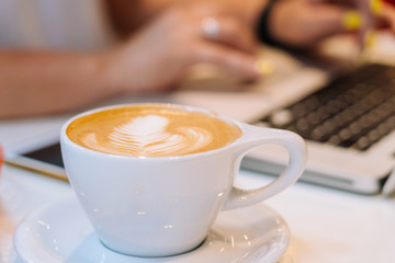 Close up cup of cappuccino coffee and hands woman sitting in office coworking and typing on her laptop keyboard computer. Desk with a phone, notebook, glasses and a cup of coffee. Business concept