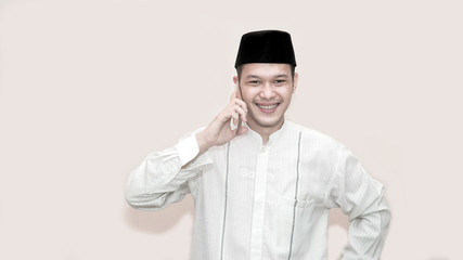 portrait of handsome asian muslim man with head cap and using smartphone taking a call and smile at camera