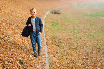 Happy boy with backpack walking in autumn park. Fashion outfit for kid. Autumn boys fashion. Cute kid enjoying fallen leaves. Happy autumn holidays