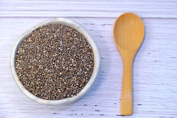 Close up Chia seeds in little ceramic bowl and an empty wooden spoon on white wooden table.