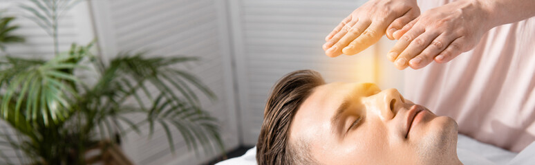 panoramic shot of healer standing near man with closed eyes and holding hands above his head