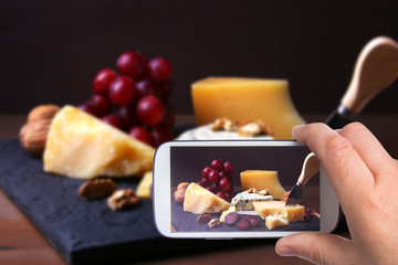hands of man with smartphone taking photo Assorted cheeses, nuts, grapes, fruits, smoked meat and a glass of wine on a serving table. Dark and Moody style.