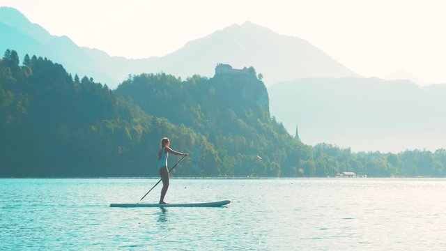 Young fitness woman in white swimwear is paddling on a SUP in the middle of the lake Bled with a castle in the background on a beautiful summer morning.