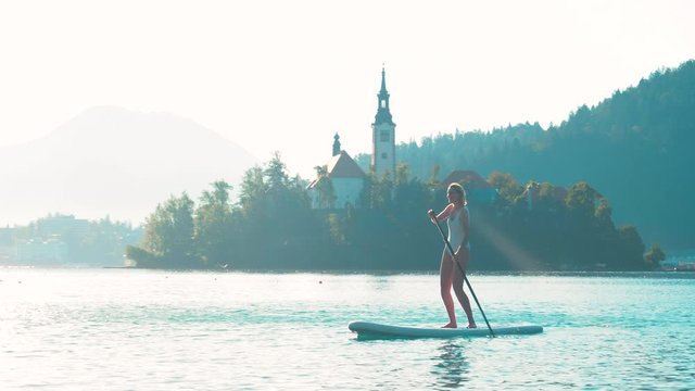 Young fitness woman in white swimwear is paddling on a SUP in the middle of the lake Bled with an island and a church in the background. 