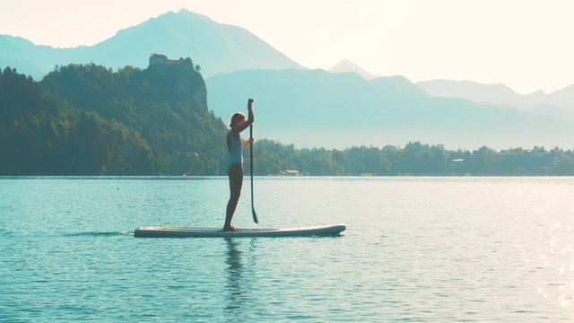 Young fitness woman is paddling on a stand-up paddle in the middle of the lake Bled with a castle and a mountain range and a sunrise in the background.