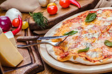 Pizza four cheeses with basil on a wooden board, wooden background decorated with vegetables and cutlery