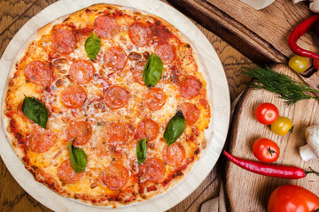 pepperoni pizza decorated with basil on a wooden board, the background is wooden decorated with vegetables and cutlery