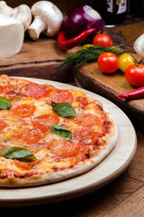 pepperoni pizza decorated with basil on a wooden board, the background is wooden decorated with vegetables and cutlery