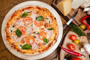 pizza with mushrooms, tomatoes, ham decorated with basil on a wooden board, the background is wooden decorated with vegetables and cutlery