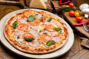 pizza with mushrooms, tomatoes, ham decorated with basil on a wooden board, the background is wooden decorated with vegetables and cutlery
