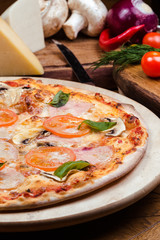 pizza with mushrooms, tomatoes, ham decorated with basil on a wooden board, the background is wooden decorated with vegetables and cutlery