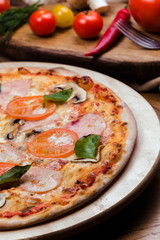 pizza with mushrooms, tomatoes, ham decorated with basil on a wooden board, the background is wooden decorated with vegetables and cutlery
