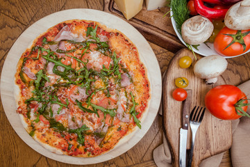 Italian pizza with ham, tomatoes, cheese and arugula on a wooden board, the background is wooden decorated with vegetables and cutlery
