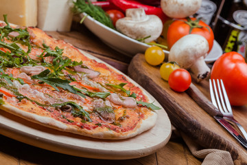 Italian pizza with ham, tomatoes, cheese and arugula on a wooden board, the background is wooden decorated with vegetables and cutlery
