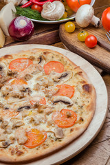 Italian pizza with chicken, tomatoes, mushrooms and cheese on a wooden board, the background is wooden decorated with vegetables and cutlery