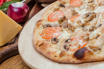 Italian pizza with chicken, tomatoes, mushrooms and cheese on a wooden board, the background is wooden decorated with vegetables and cutlery