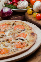 Italian pizza with chicken, tomatoes, mushrooms and cheese on a wooden board, the background is wooden decorated with vegetables and cutlery