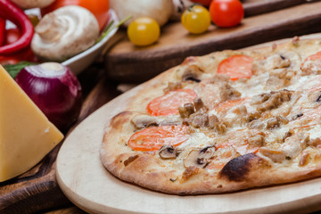 Italian pizza with chicken, tomatoes, mushrooms and cheese on a wooden board, the background is wooden decorated with vegetables and cutlery