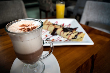 cappuccino served in a elegant cup accompanied by an orange juice and a pancake filled with dulce de leche and chocolate