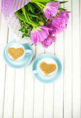 Two cups of cappuccino with a heart shaped symbol and purple tulips on a wooden background, close-up