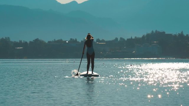 A handheld shot of a young female in a white swimwear paddling on a stand-up paddle in the middle of the lake with a sun rising in the background.