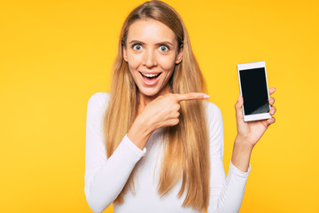 Fototapeta premium Did you see this one? Excited beautiful young blonde girl while she pointing on blank screen of smart phone.
