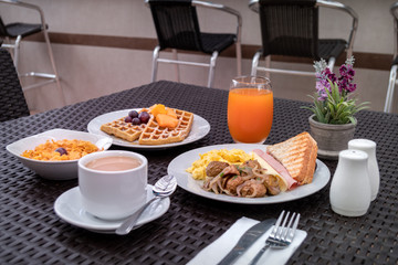 Breakfast consisting of a coffee with milk, sandwich with cheese and cold meats, garnishes, cereals with yogurt, waffles with some fruits and an orange juice