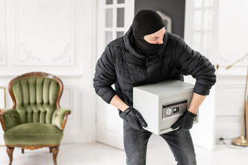 Thief with black balaclava stealing modern Electronic safe box. The burglar commits a crime in...