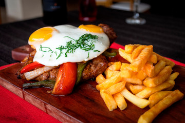 Juicy meat accompanied with sauteed vegetables fried egg and french fries, all served on a wooden board