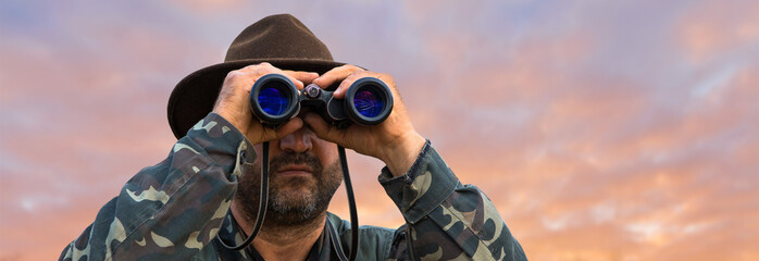 A hunter in a hat with binoculars looks out for prey against the background of the sunset.