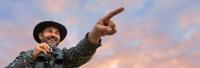 A hunter in a hat with binoculars looks out for prey against the background of the sunset.