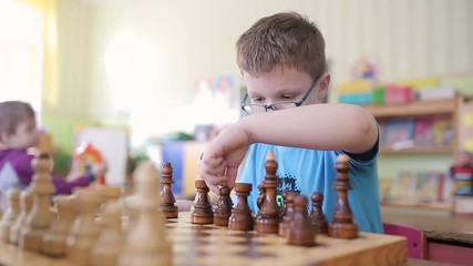 A boy with glasses plays chess. He is sitting at the table playing and smiling emotionally. Rearranges pieces on a chessboard. He wants to checkmate and win.