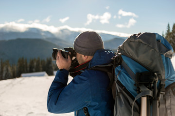 Fototapeta premium Hiker man tourist photographer in warm clothing with backpack and camera taking picture of snowy valley and woody mountain peaks landscape under blue sky on sunny winter cold day.