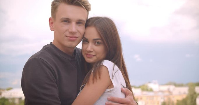 Closeup portrait of young cute caucasian couple hugging looking at camera standing on the balcony of their house with beautiful landscape on the background