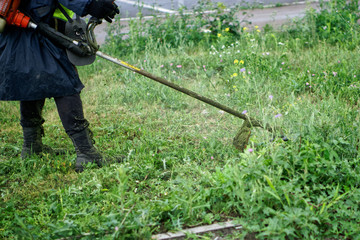 A man mows weeds and grass on a city lawn using a hand-held lawn mower.