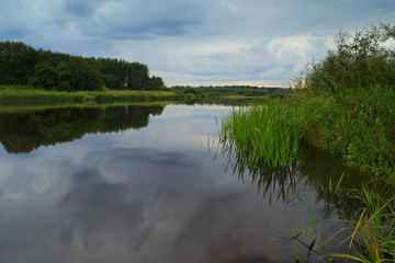 Fototapeta na wymiar July sunset before the rain over the river, with heavy, rainy clouds reflected in the mirror river and a strip of sunlight over the forest.