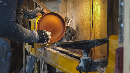 Woodworker grinds the workpiece by sawdust. Carpenter completes work at lathe. Making beautiful wooden dishes handmade. Builder works with workpiece in workshop. Creation unusual dishes.
