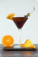 Dark orange drink in a glass. Ice cubes and lemon slices. Green and black olives. Vermouth and gin.