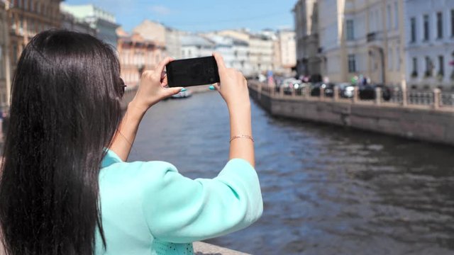 Smiling travel woman enjoying shooting video of water canal and cityscape from embankment in European city