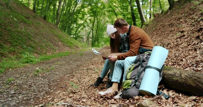 Male and female backpacker hikers sitting on log in forest and checking direction on map, trakking