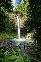 A young man walking on a path that leads to La Fortuna waterfall with a backpack and a hat,...
