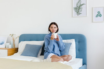 Girl in a blue sweater in interior Hygge style with a cup in hand sits on the bed